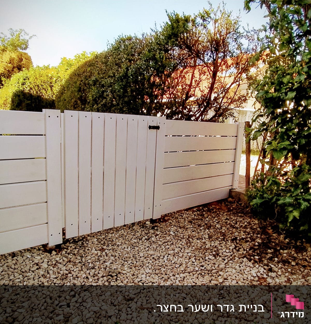 גדר עץ לבנה על חצץ ליד צמחייה ירוקה